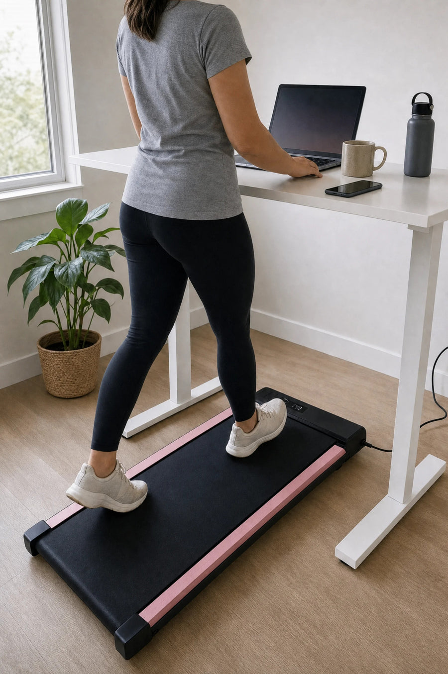 Woman walking on the pink under-desk walking pad (black belt with soft-pink side rails) at a white standing desk while working on her laptop