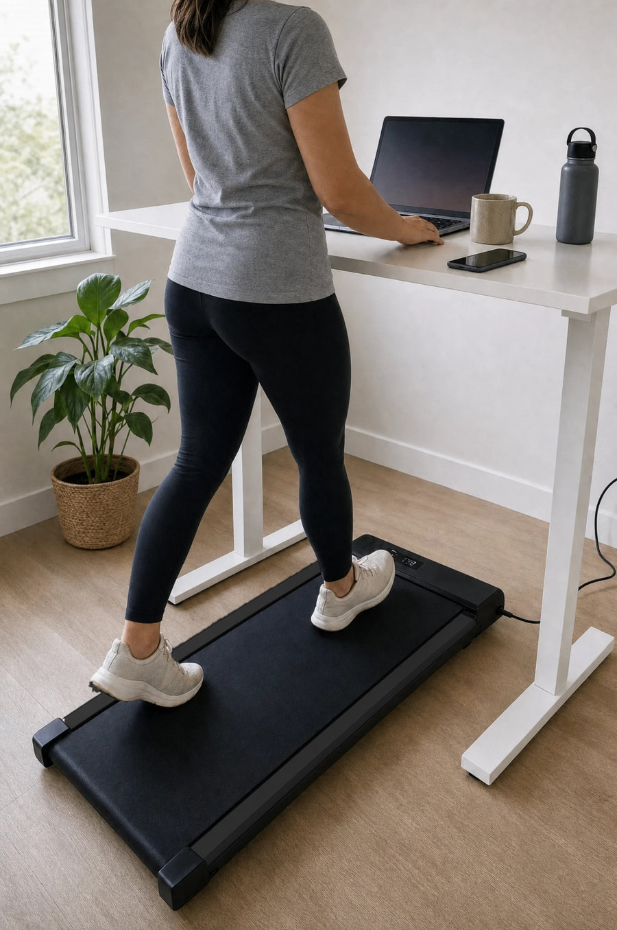Woman walking on the black under-desk walking pad at a white standing desk while working on her laptop