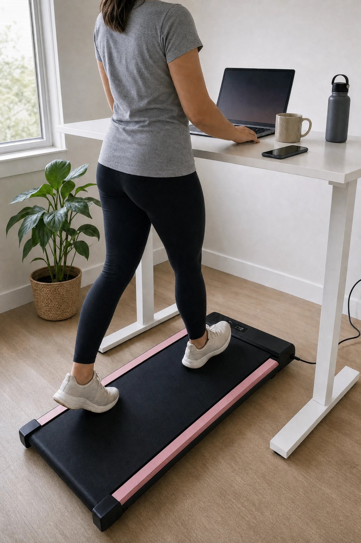 Woman walking on the pink under-desk walking pad (black belt with soft-pink side rails) at a white standing desk while working on her laptop
